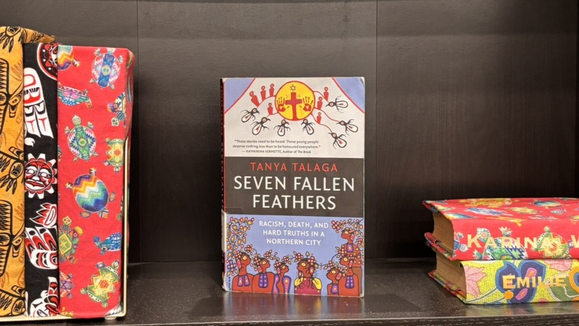 A copy of Tanya Talaga’s Seven Fallen Feathers stands upright on a shelf in King’s library, surrounded by books wrapped in colourful patterned fabric.